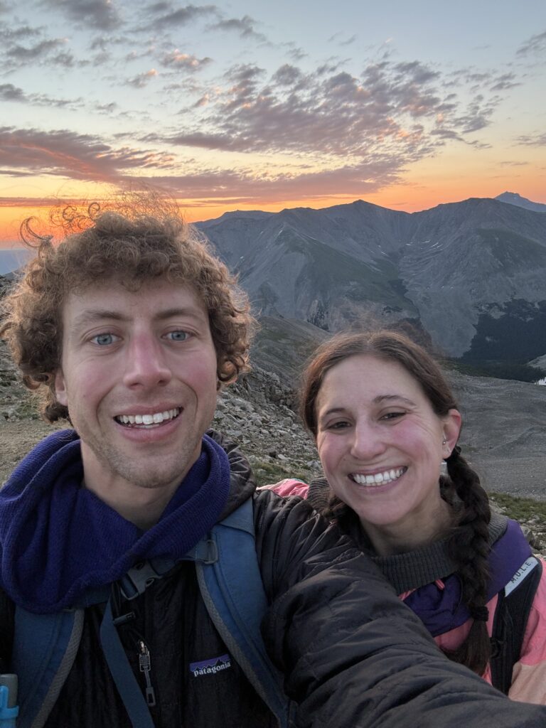 Jon and Lai smiling in a selfie with the sunrise over the mountains in the background toward the top of Huron Peak.