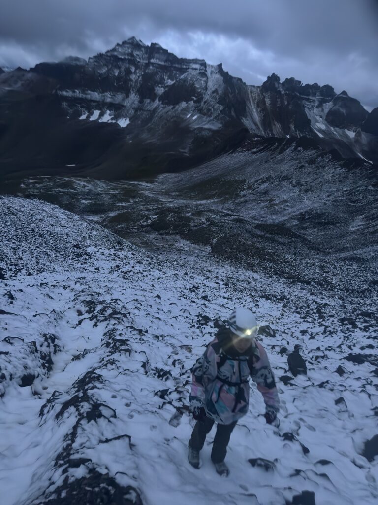 Lai hiking in the snow with a headlamp on in the dim lighting. Rugged peaks are seen in the background