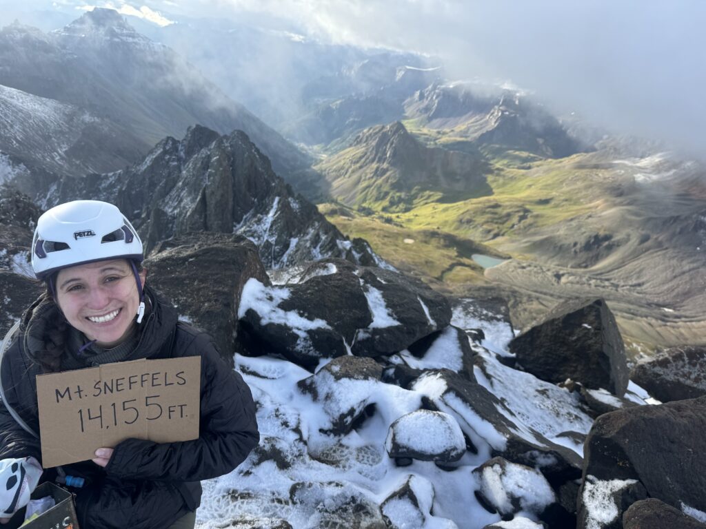 Lai at the top of Mount Sneffels with a cardboard sign with details of the peak. Greenery and a lake are in the bacground.