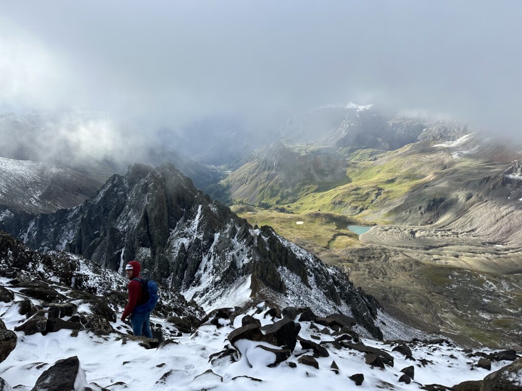 Jon toward the top of Mount Sneffels among rugged snow capped peaks, with green mountains and a lake in the background