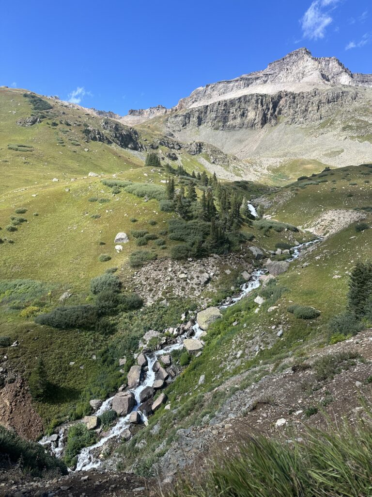 A waterfall trickling down green mountains with a gray mountain and a blue sky